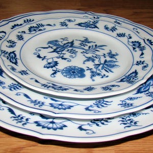 May include: Three white porcelain plates with a blue floral design are stacked on a wooden surface. The plates have a scalloped edge and a detailed blue floral pattern. The plates are of different sizes.