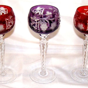 May include: Three stemmed wine glasses with intricate cut glass designs. Two glasses are ruby red, and one is purple. The stems are clear, with a twisted design, and the bases are circular.