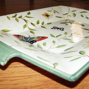 May include: A rectangular serving platter with a white surface, adorned with painted butterflies and green foliage. The platter has a light green border and a handle-like extension on one side. The butterflies are in various colors.