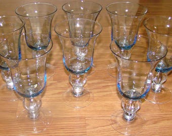 May include: Set of eight clear glass goblets with a flared rim and a blue accent. Each goblet has a round base and a decorative stem. The glasses are arranged on a wooden surface.