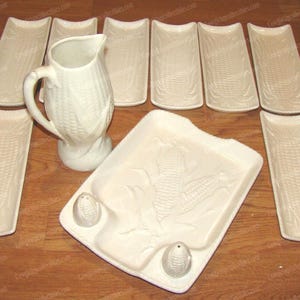 May include: A cream-colored ceramic dinnerware set featuring a corn-themed embossed pattern. The set includes a pitcher, a large rectangular serving platter with two integrated corn-shaped shakers, and seven long, narrow rectangular corn-shaped plates.