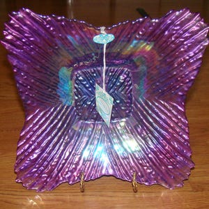 May include: A decorative iridescent purple glass bowl with a ruffled edge. The bowl has a square center and a tag attached with a string. The bowl is displayed on a gold stand.