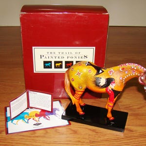May include: A decorative horse figurine from "The Trail of Painted Ponies" collection. The horse is painted with yellow, orange, and red colors, and features a Native American design. It stands on a black base, with a red box and informational cards in the background.