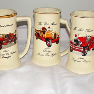 May include: Five cream-colored ceramic beer steins with gold rims and handles. Each stein features a vintage fire truck illustration and the text "The Last Alarm" with a year and model name. The steins are arranged on a white surface.