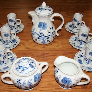 May include: A white porcelain tea set with blue floral designs. The set includes a teapot, creamer, sugar bowl, and six teacups with saucers. The tea set is arranged on a wooden surface.