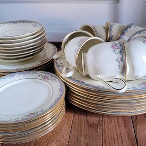 May include: A collection of vintage porcelain dishware, including plates, saucers, and teacups. The set features a white base with a blue floral border and gold trim. The plates and saucers are stacked, and the teacups are arranged on top of the saucers.