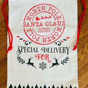 May include: A white canvas Santa Claus gift bag with red drawstring closure. The bag features a red circular stamp that reads "North Pole Santa Claus Express Delivery." Below the stamp, the words "Special Delivery For" are printed in black.