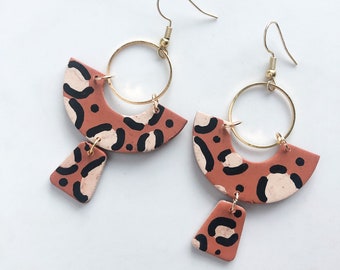May include: A pair of handmade earrings. The earrings have a terracotta-coloured, semi-circular top with black and cream-coloured spots, connected to a gold hoop and a smaller, matching pendant. The earrings have gold-coloured ear wires. A unique design.