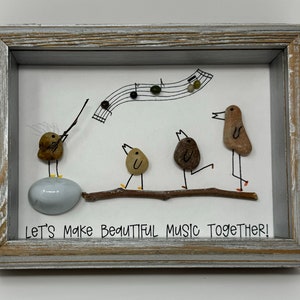 Könnte beinhalten: Gerahmtes Kunstwerk mit vier kleinen Vögeln aus bemalten Steinen, wobei ein Vogel ein Musikinstrument spielt. Die Vögel sitzen auf einem Ast mit darüber gezeichneten Noten. Das Kunstwerk trägt unten den Text "Let's Make Beautiful Music Together!"
