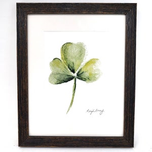 Puede incluir: Pintura a la acuarela de un tr&eacute;bol verde de tres hojas, firmada "Leigh Barry", enmarcada en un marco de madera marr&oacute;n oscuro. El tr&eacute;bol est&aacute; centrado sobre un fondo blanco.
