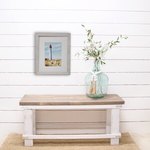 Puede incluir: Una pintura de acuarela enmarcada de un faro en una costa rocosa cuelga de una pared de tablones blancos. Un jarr&oacute;n de cristal transparente con tallos verdes y cuentas blancas se encuentra sobre un banco de madera con patas blancas.