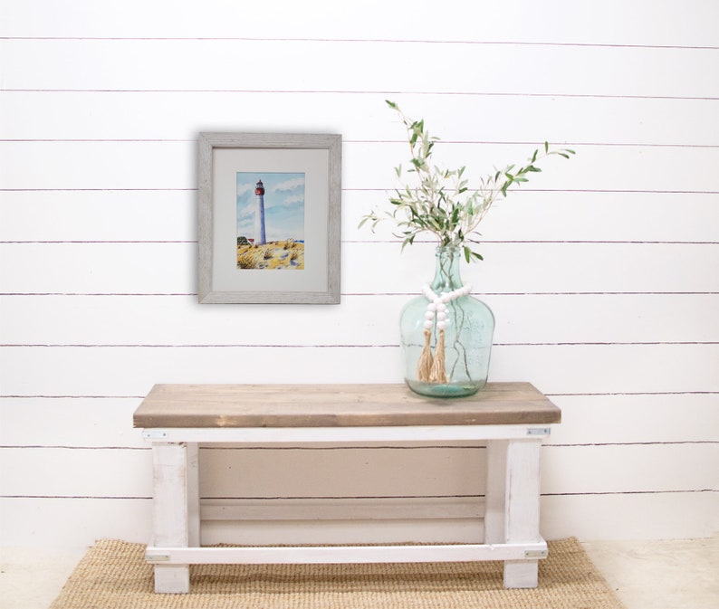 Puede incluir: Una pintura de acuarela enmarcada de un faro en una costa rocosa cuelga de una pared de tablones blancos. Un jarr&oacute;n de cristal transparente con tallos verdes y cuentas blancas se encuentra sobre un banco de madera con patas blancas.