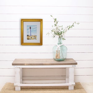 Puede incluir: Una pintura de acuarela enmarcada de un faro con un marco dorado. La pintura se exhibe en una pared blanca sobre un banco de madera. Un jarr&oacute;n de cristal con vegetaci&oacute;n y cuentas decorativas se encuentra en el banco.
