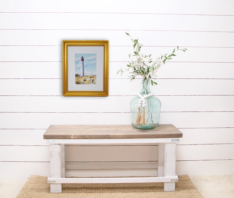 Puede incluir: Una pintura de acuarela enmarcada de un faro con un marco dorado. La pintura se exhibe en una pared blanca sobre un banco de madera. Un jarr&oacute;n de cristal con vegetaci&oacute;n y cuentas decorativas se encuentra en el banco.