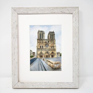 May include: Watercolour painting of Notre Dame Cathedral in Paris, France, framed in a white, distressed wood frame. The artwork depicts the cathedral's facade with its iconic towers, rose window, and detailed architectural elements. The painting is set against a light blue sky.