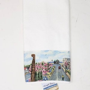 May include: White hand towel with a watercolor painting of a street scene. The artwork depicts a road lined with pink flowering trees, buildings, and a wooden totem. The towel has a tag that reads "Leigh Barry Watercolors, Maryland".