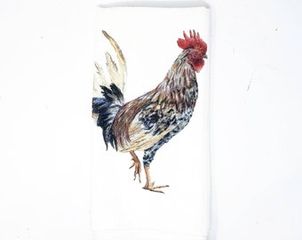 May include: White hand towel featuring a detailed watercolor illustration of a rooster. The rooster has brown, black, and white feathers, a red comb, and a yellow beak. The towel is folded and has a stitched edge.