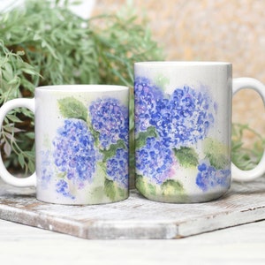 May include: Two white ceramic mugs with a watercolor design of blue and purple hydrangeas. The mugs have a curved handle and a white background. The flowers have green leaves and are painted in a soft, artistic style.