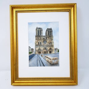 May include: A framed watercolour painting of Notre Dame Cathedral in Paris, France. The artwork is set within a gold-coloured ornate frame. The painting depicts the cathedral's facade, including its towers, rose window, and architectural details, against a light blue sky.