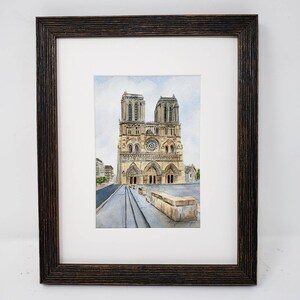 May include: Watercolour painting of Notre Dame Cathedral in Paris, France, framed in a dark brown wooden frame. The artwork depicts the cathedral's facade with its iconic towers, rose window, and architectural details. The painting is set against a light blue sky.
