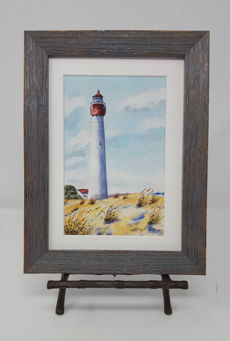 Puede incluir: Pintura a la acuarela de un faro blanco alto con una parte superior roja, sobre un cielo azul con nubes. La obra de arte est&aacute; enmarcada en un marco de madera gris y se exhibe en un soporte decorativo. La escena incluye dunas de arena y vegetaci&oacute;n escasa.