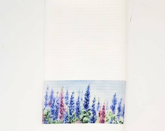 May include: White hand towel with a watercolor design of purple and blue lupine flowers. The floral design is at the bottom of the towel, with a light blue background. The towel has a textured surface.