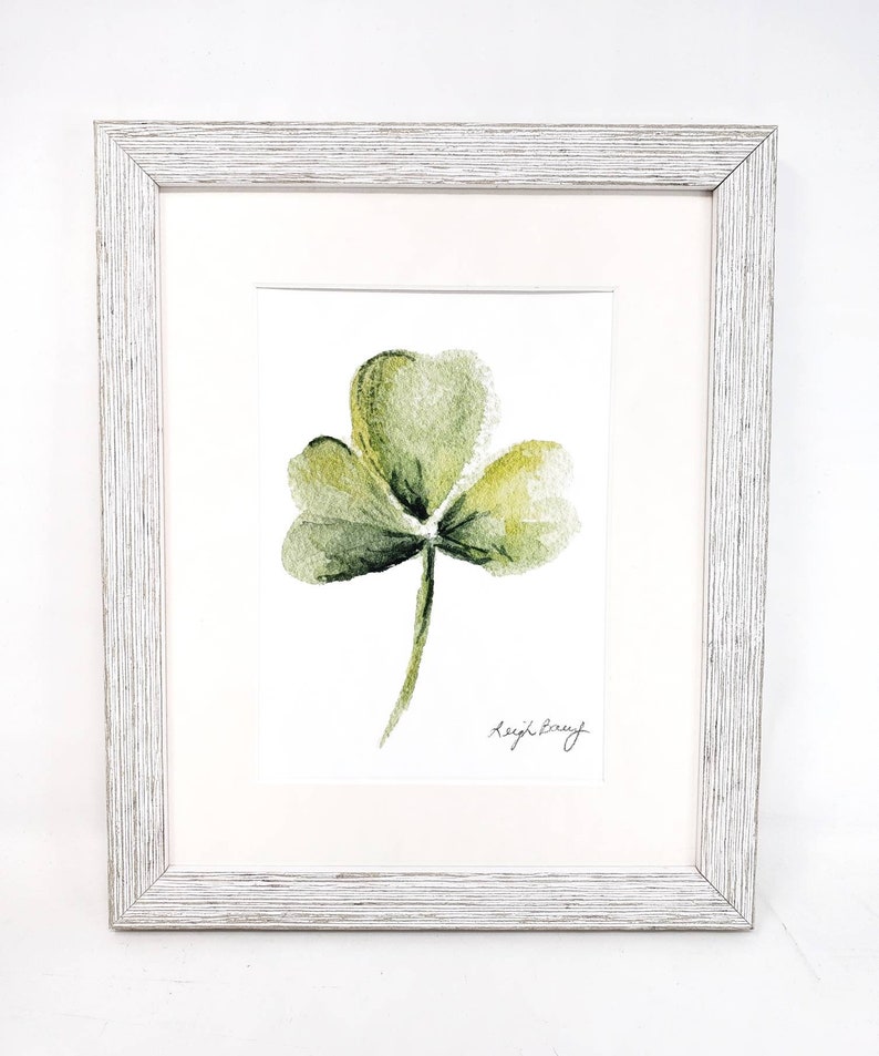 Puede incluir: Una pintura de acuarela enmarcada de un tr&eacute;bol de tres hojas. La obra presenta tonos de verde y amarillo, con un passe-partout blanco y un marco de madera blanca envejecida. La firma del artista, "Leigh Barry", es visible en la esquina inferior derecha.