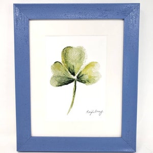 Puede incluir: Una pintura de acuarela enmarcada de un tr&eacute;bol verde con un marco de madera azul. La obra de arte presenta un tr&eacute;bol de tres hojas con sombreado delicado y la firma del artista.