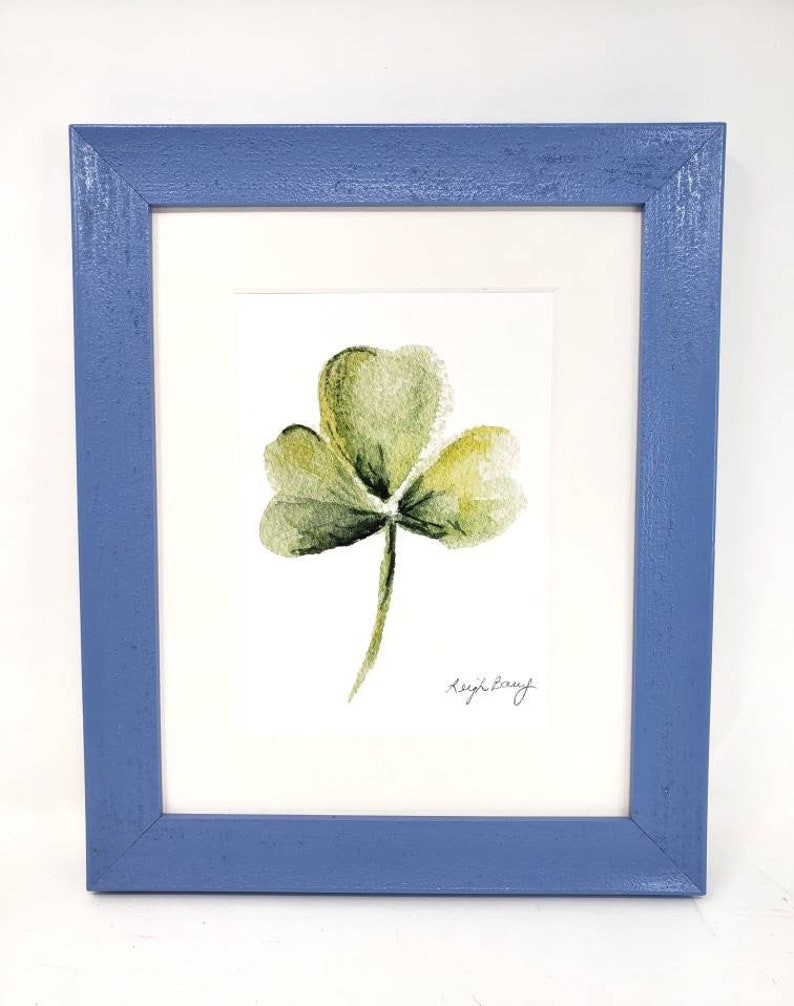 Puede incluir: Una pintura de acuarela enmarcada de un tr&eacute;bol verde con un marco de madera azul. La obra de arte presenta un tr&eacute;bol de tres hojas con sombreado delicado y la firma del artista.