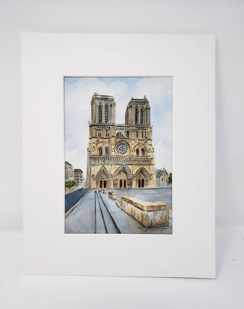 May include: Watercolour painting of Notre Dame Cathedral in Paris, France, presented in a white mount. The artwork shows the cathedral's facade with its iconic towers, rose window, and arched entrance. The painting uses muted tones of beige, grey, and blue.
