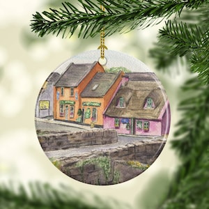 May include: Round Christmas ornament featuring a watercolor painting of a quaint village scene. The artwork depicts colorful buildings with thatched roofs and a stone bridge. The ornament is suspended by a gold string against a blurred background.