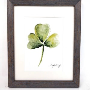 Puede incluir: Pintura de acuarela enmarcada de un tr&eacute;bol verde de tres hojas. La obra de arte est&aacute; firmada "Leigh Barry" en la esquina inferior derecha. La pintura est&aacute; enmarcada en un marco de madera marr&oacute;n oscuro.