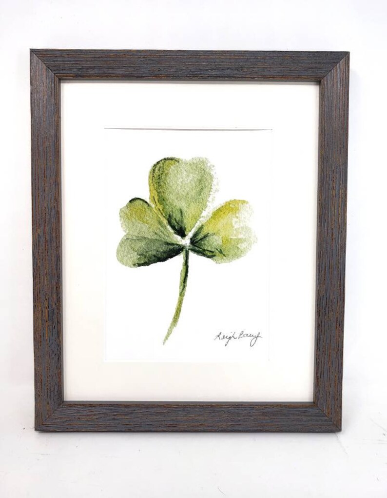 Puede incluir: Pintura de acuarela enmarcada de un tr&eacute;bol verde de tres hojas. La obra de arte est&aacute; firmada "Leigh Barry" en la esquina inferior derecha. La pintura est&aacute; enmarcada en un marco de madera marr&oacute;n oscuro.