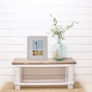 Puede incluir: Una pintura de acuarela enmarcada de un faro con la parte superior roja, sobre un cielo azul y terreno arenoso. La pintura se exhibe en un banco de madera r&uacute;stico, junto a un jarr&oacute;n de vidrio transparente con vegetaci&oacute;n y cuentas decorativas.