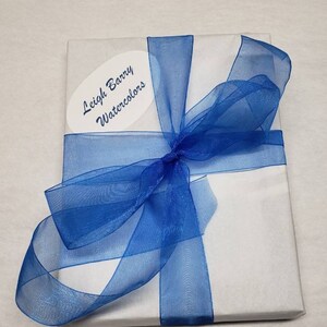Puede incluir: Un paquete envuelto en papel blanco, atado con una cinta azul transparente. Una etiqueta ovalada con el texto "Leigh Barry Watercolors" est&aacute; adherida. La presentaci&oacute;n sugiere un regalo o una compra especial.