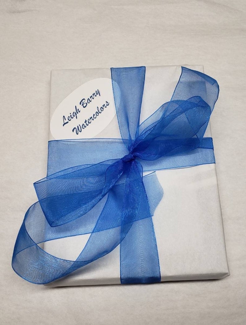 Puede incluir: Un paquete envuelto en papel blanco, atado con una cinta azul transparente. Una etiqueta ovalada con el texto "Leigh Barry Watercolors" est&aacute; adherida. La presentaci&oacute;n sugiere un regalo o una compra especial.