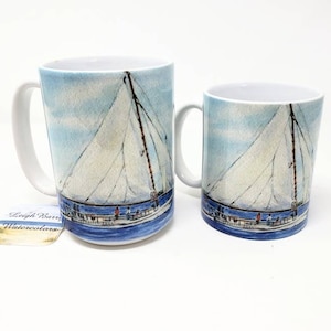 May include: Two white ceramic mugs with a watercolor painting of a sailboat. The design features a blue sky, white sails, and a blue hull. The mugs have white handles and the text "Leigh Barn Watercolors" on a small tag.