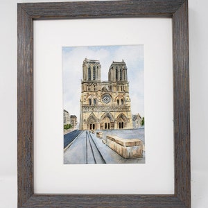 May include: Watercolour painting of Notre Dame Cathedral in Paris, France, in a grey wooden frame. The artwork shows the cathedral's facade with its rose window and towers, against a cloudy sky. The painting is on white mount board.