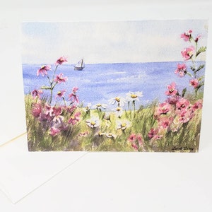 Puede incluir: Tarjeta de felicitaci&oacute;n de acuarela con una escena costera. Flores silvestres rosas y blancas en primer plano, con un velero en el agua azul bajo un cielo azul claro. Incluye un sobre blanco.