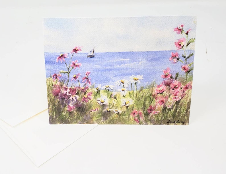 Puede incluir: Tarjeta de felicitaci&oacute;n de acuarela con una escena costera. Flores silvestres rosas y blancas en primer plano, con un velero en el agua azul bajo un cielo azul claro. Incluye un sobre blanco.