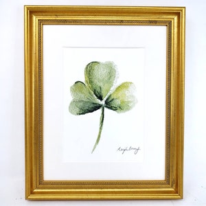 Puede incluir: Pintura de acuarela enmarcada de un tr&eacute;bol verde de tres hojas. La obra de arte est&aacute; sobre un fondo blanco y enmarcada en un marco dorado y ornamentado. La firma del artista es visible en la esquina inferior derecha.