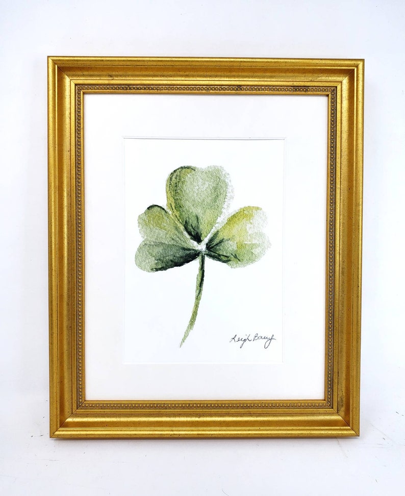 Puede incluir: Pintura de acuarela enmarcada de un tr&eacute;bol verde de tres hojas. La obra de arte est&aacute; sobre un fondo blanco y enmarcada en un marco dorado y ornamentado. La firma del artista es visible en la esquina inferior derecha.