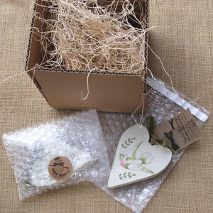 May include: A white heart-shaped wooden ornament with a green bird and leaves painted on it. The ornament is packaged in a bubble wrap envelope with a small tag that reads "Smile Darling, Everything Will Be Alright".