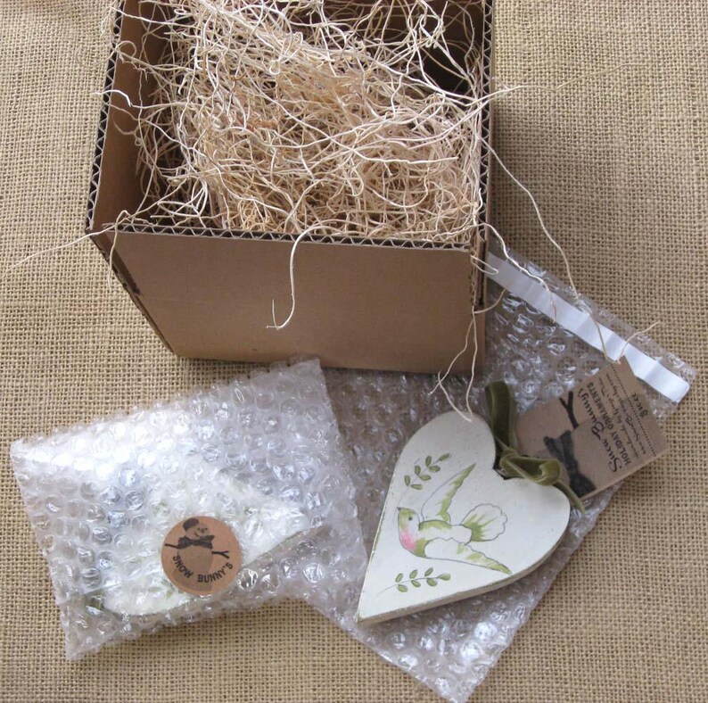 May include: A white heart-shaped wooden ornament with a green bird and leaves painted on it. The ornament is packaged in a bubble wrap envelope with a small tag that reads "Smile Darling, Everything Will Be Alright".