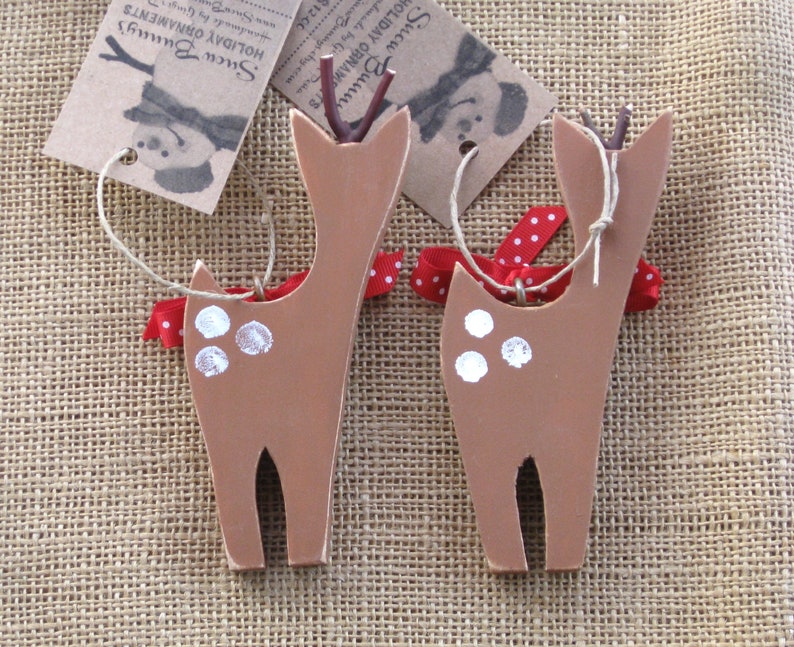 May include: Two brown wooden deer ornaments with white spots and red and white polka dot ribbon. The ornaments have twine attached for hanging.