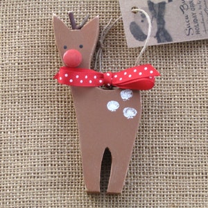 May include: A wooden reindeer ornament with a red nose, white spots, and a red and white polka dot bow. The ornament is hanging from a piece of twine.