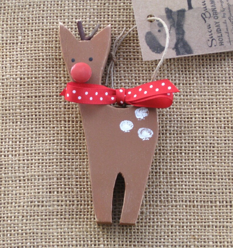 May include: A wooden reindeer ornament with a red nose, white spots, and a red and white polka dot bow. The ornament is hanging from a piece of twine.