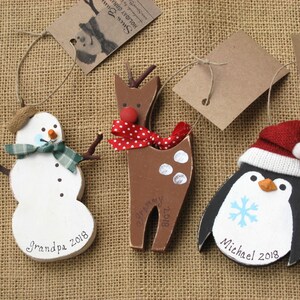 May include: Three wooden Christmas ornaments, a snowman, a reindeer, and a penguin. The snowman is white with a green scarf and a brown hat. The reindeer is brown with white spots and a red and white polka dot scarf. The penguin is black with a white belly and a red and white hat. Each ornament has a name and the year 2018 painted on it.
