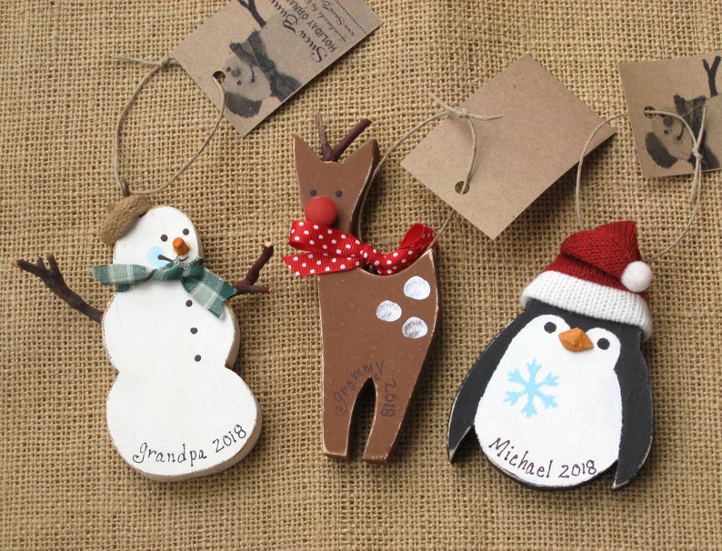 May include: Three wooden Christmas ornaments, a snowman, a reindeer, and a penguin. The snowman is white with a green scarf and a brown hat. The reindeer is brown with white spots and a red and white polka dot scarf. The penguin is black with a white belly and a red and white hat. Each ornament has a name and the year 2018 painted on it.