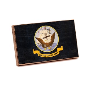 May include: A rectangular wooden plaque featuring the United States Navy seal on a dark blue background. The seal displays a bald eagle with wings spread, a ship, and an anchor, with a yellow banner below reading UNITED STATES NAVY.